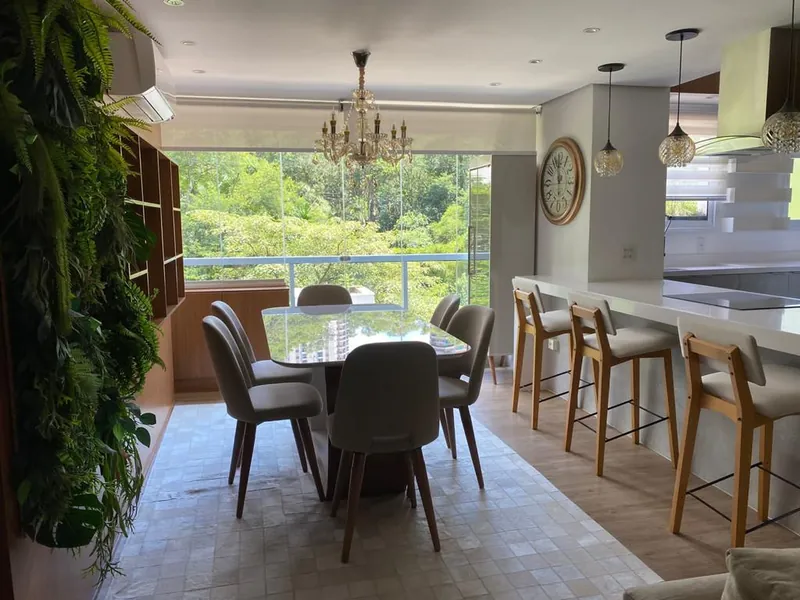 Sala de jantar com mesa de vidro, lustre de cristal, bancada com banquetas e vista para a mata