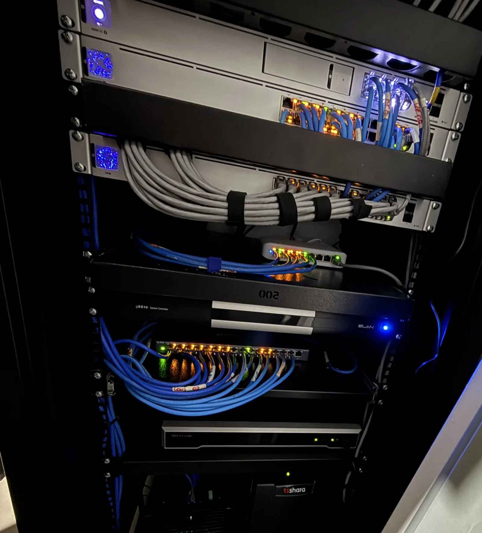 Rack de rede com switches, patch panels e cabos azuis organizados, com LEDs acesos